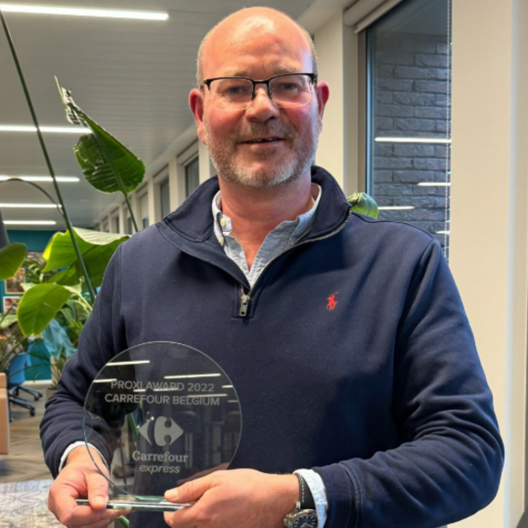 Man holding a clear award plaque indoors, standing in front of plants and windows.