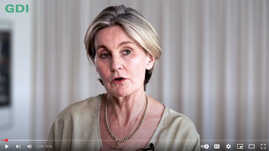 Woman speaking on camera in a softly lit indoor setting, wearing a beige top and necklace.