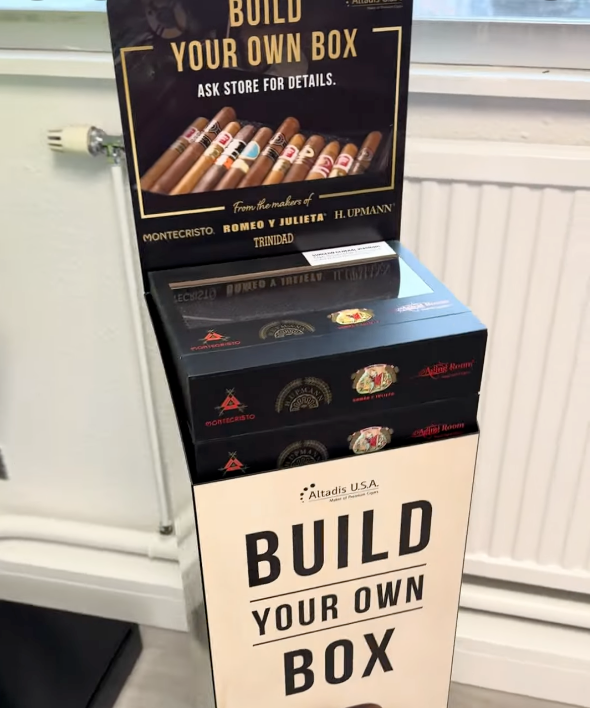 Stack of black and beige “Build Your Own Box” product boxes in a shop by a radiator.