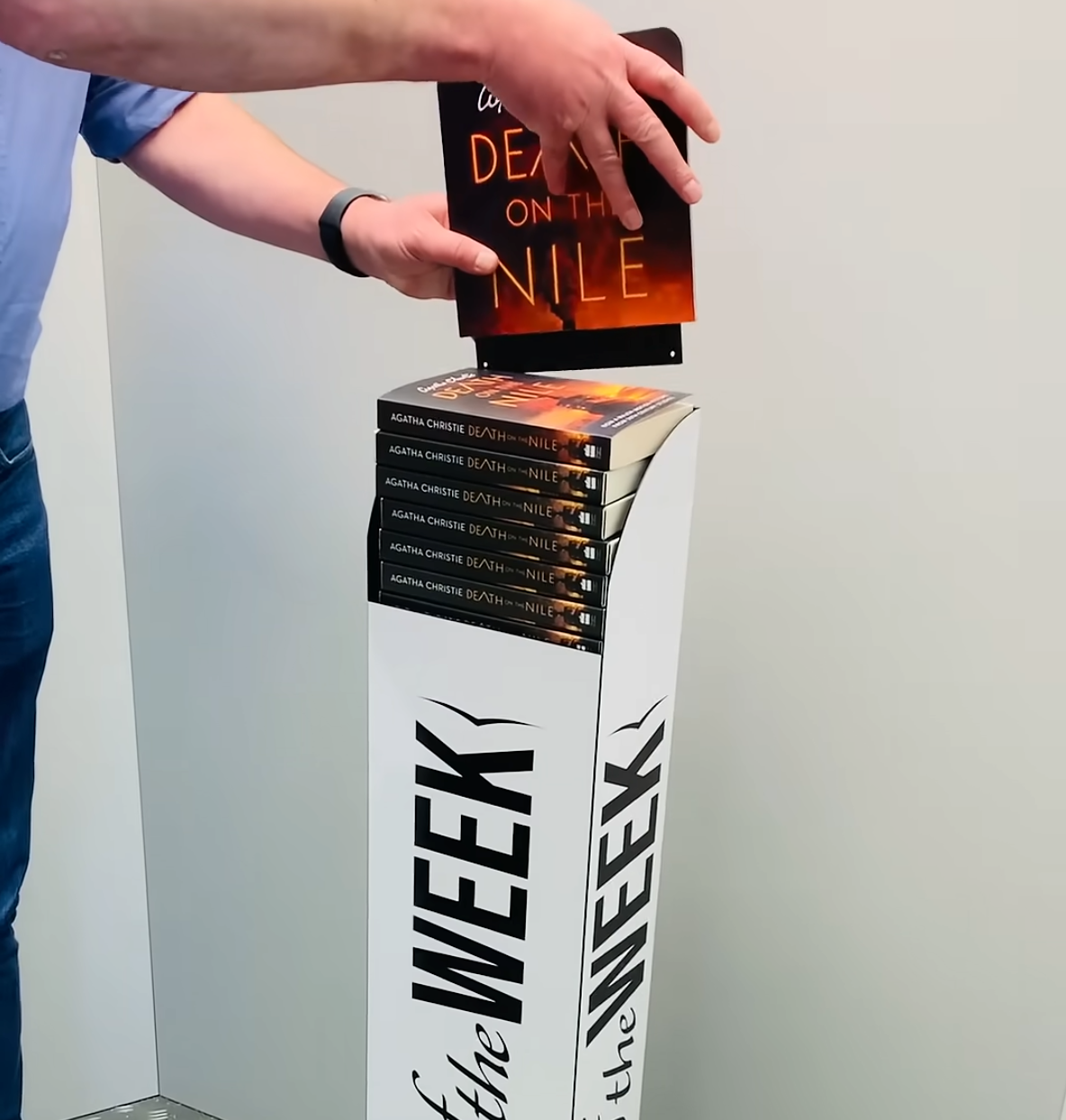 Person stacking black-and-orange books on a white WEEK display tower