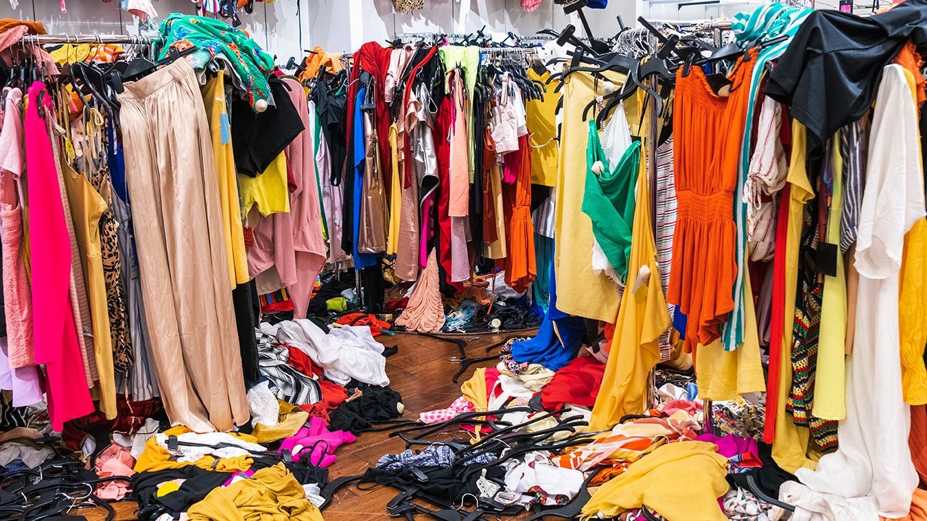 Crowded clothing market stall filled with colorful hanging garments and scattered clothes on the floor
