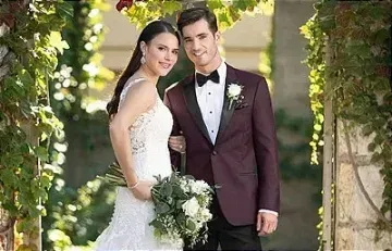 Bride and groom posing outdoors under a floral arch, smiling in wedding attire