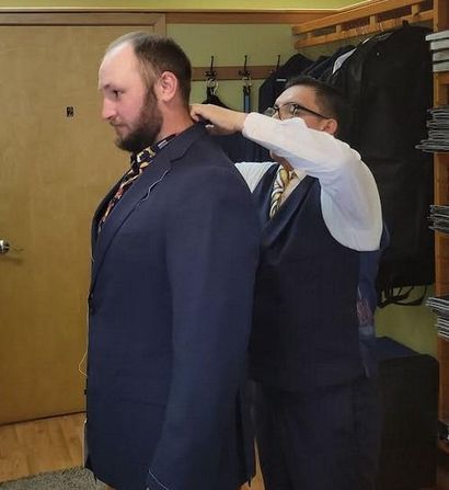 Tailor fitting a man's navy suit jacket, adjusting the collar in a fitting room