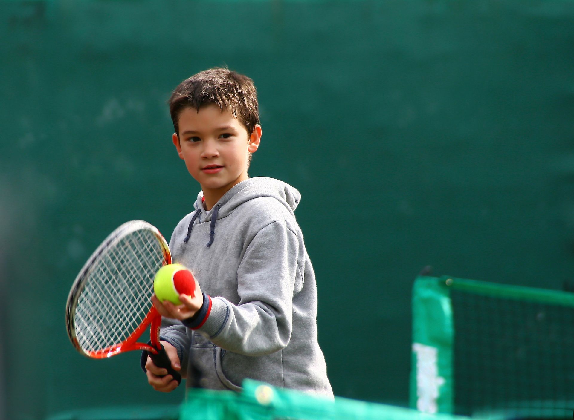 Giovane tennista con racchetta e palla su un campo, con una rete verde sullo sfondo.