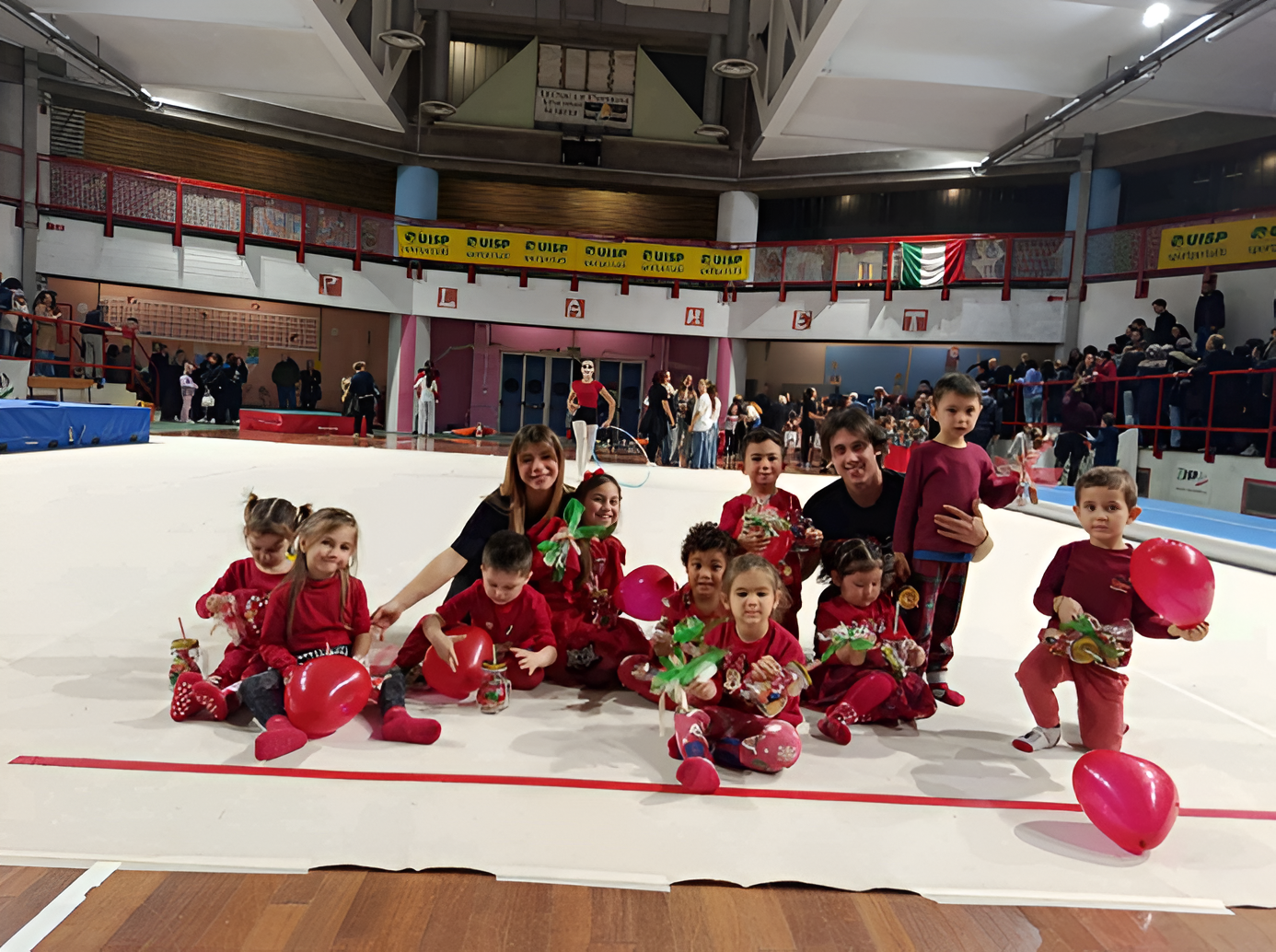 Bambini in costume rosso posano su una pista di pattinaggio al coperto con palloncini rossi, mentre gli spettatori sugli spalti si trovano alle loro spalle.