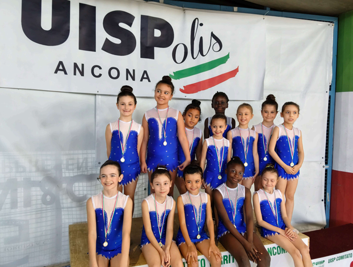 Nove ginnaste in body blu alzano le braccia su un podio, indossando le medaglie vinte durante una competizione indoor.