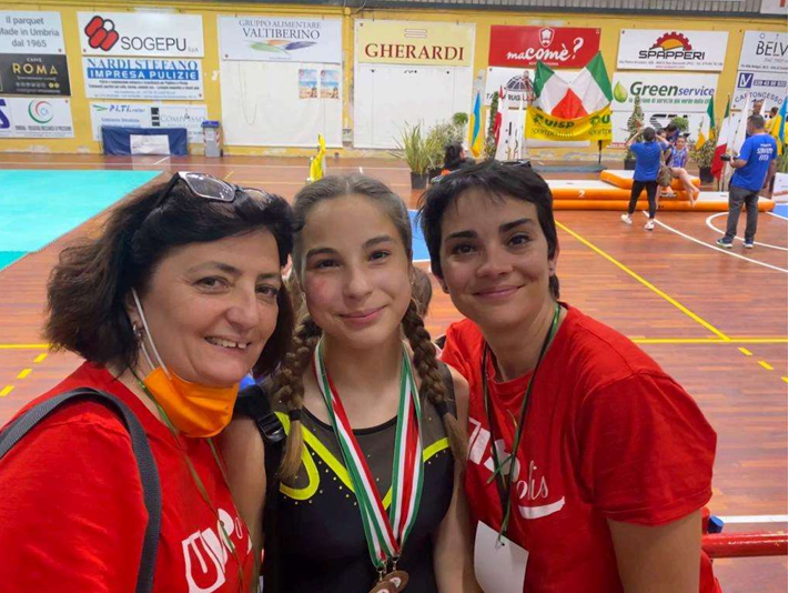 Tre atleti sorridenti con magliette rosse e medaglie, in posa in un palazzetto dello sport durante una gara di pallavolo.