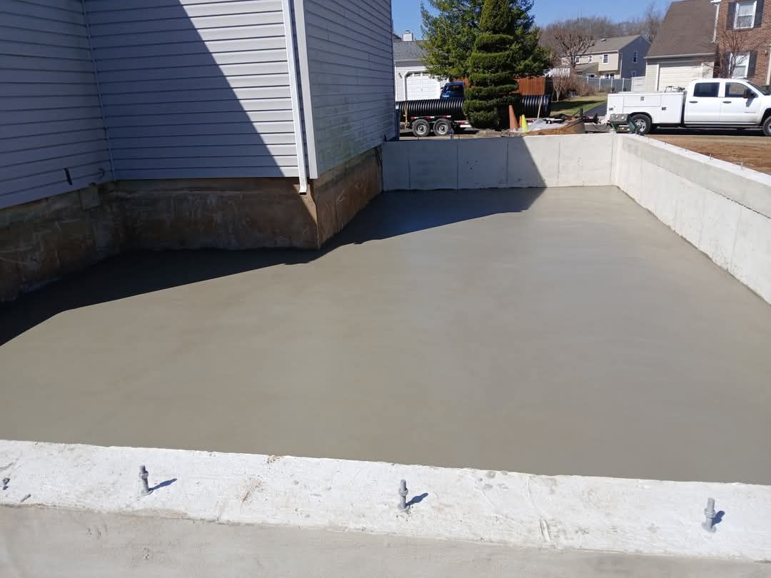 Freshly poured concrete driveway beside a house with a low retaining wall and parked vehicles in the distance