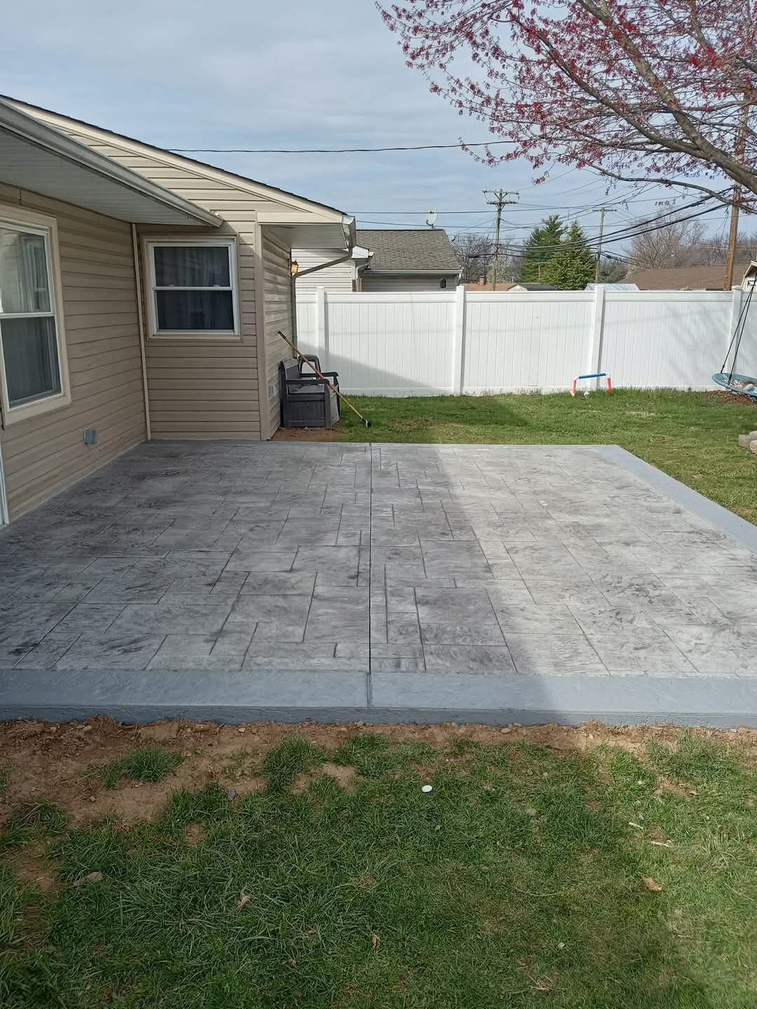 Back patio with gray pavers, grass border, white fence, and a house exterior.