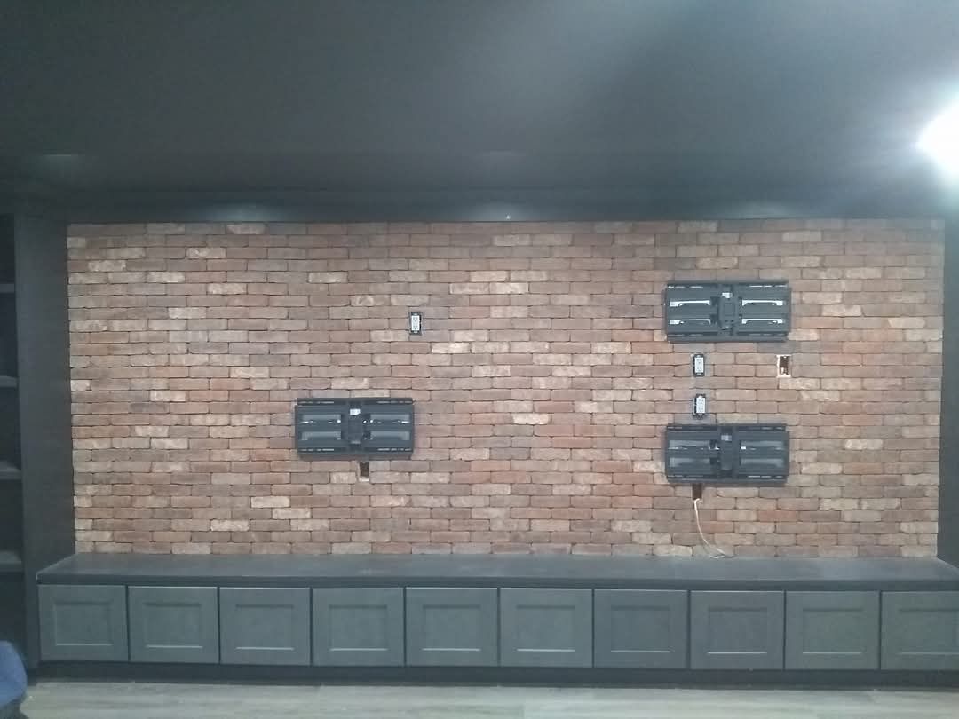 Brick wall with three mounted displays above a gray cabinet in a dim room