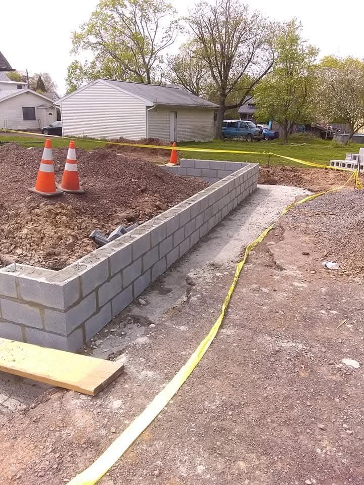 Construction site with cinder block retaining wall, orange cones, and yellow caution tape in a yard.