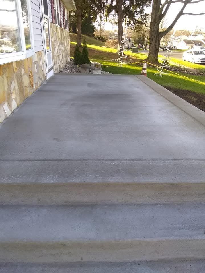 Concrete ramp beside a stone house, leading to a sunlit yard and trees