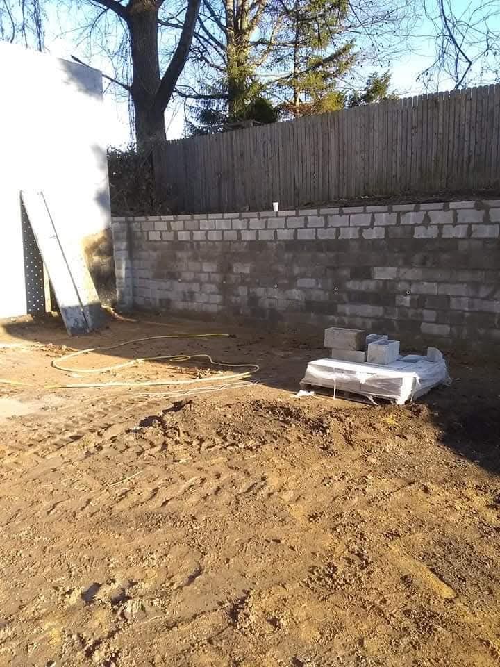 Dirt yard beside a stone wall, with stacked boards and a white pallet in sunlight.