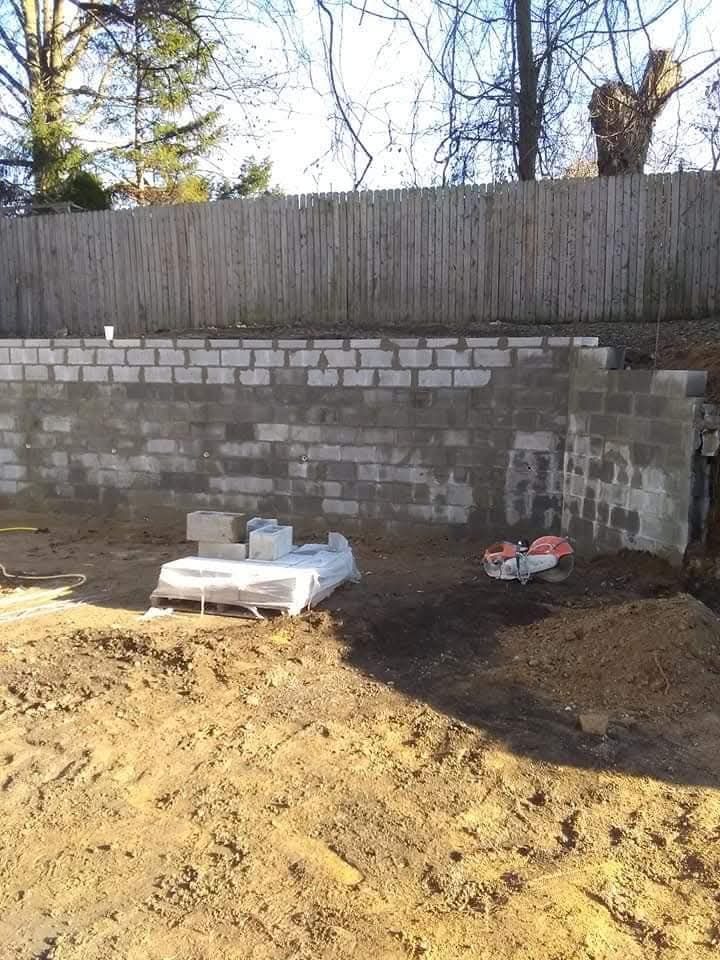 Backyard with cinderblock retaining wall, wooden fence, and construction materials on dirt ground