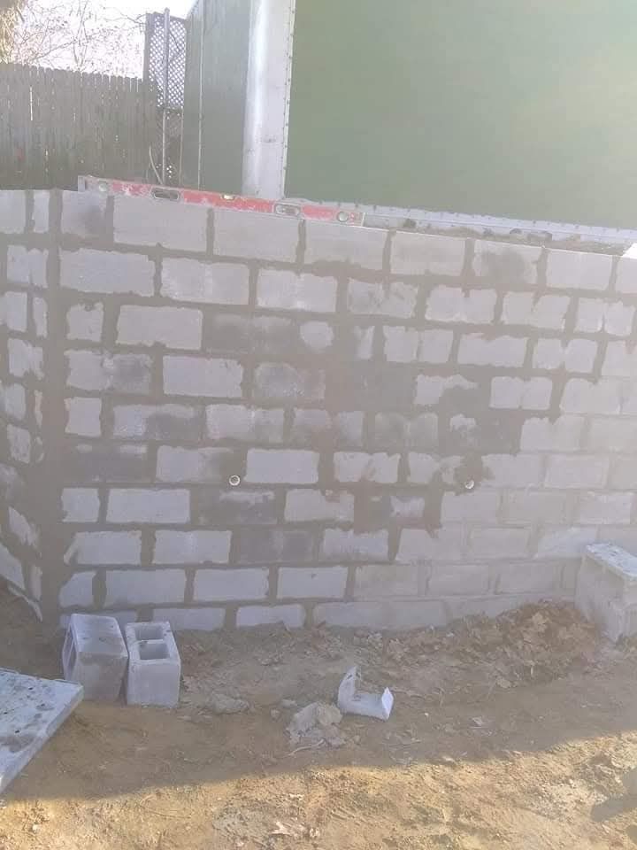 Partially built cinder block wall in a dusty outdoor construction area, with cinder blocks on the ground.