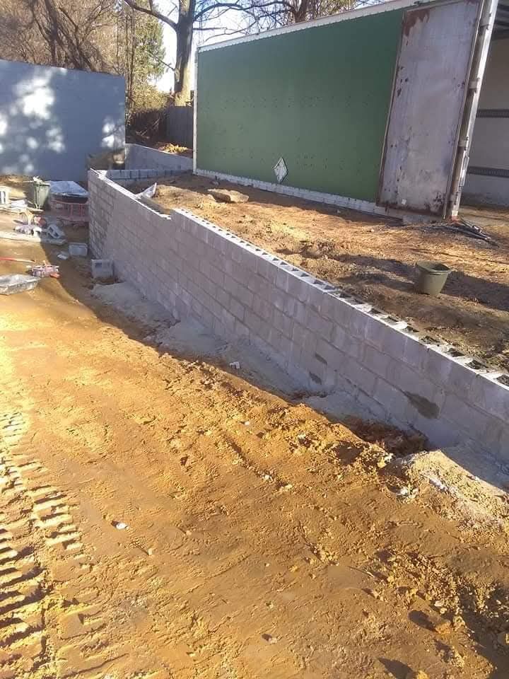 Damaged roadside retaining wall beside a dirt path and a green fence panel in bright sunlight