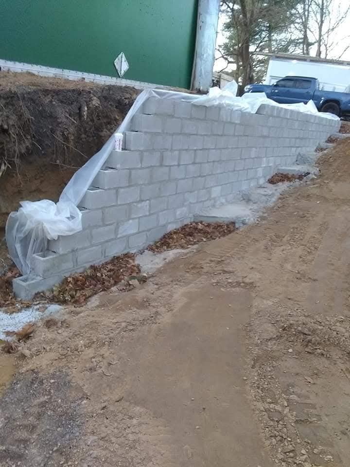 A white cinder block retaining wall beside a dirt road, with a green building and snow nearby.