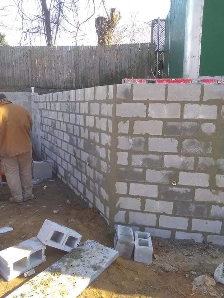 Partially built cinder block wall outdoors, with construction debris and a worker standing at left
