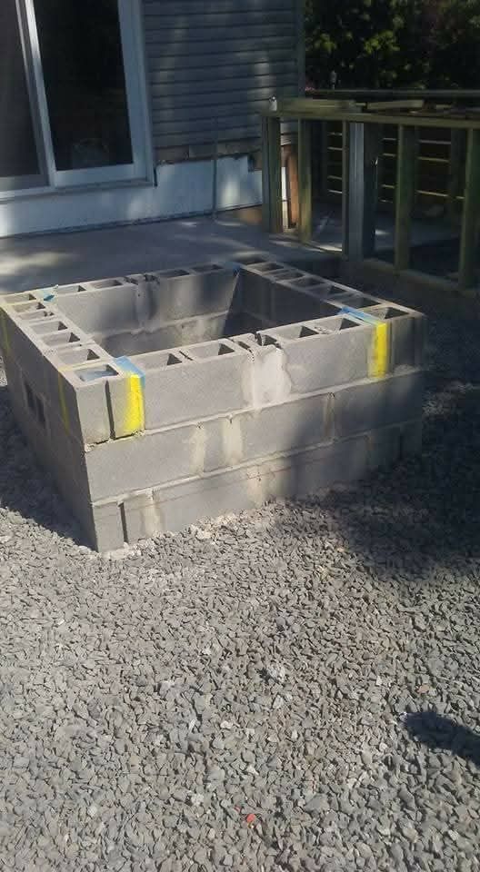 Partially built cinder block fire pit on gravel beside a house and wooden deck railing