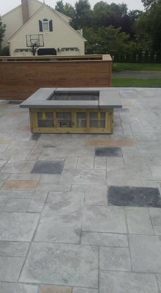 Outdoor memorial with low stone seating and a brick wall in a paved plaza, with a house and trees beyond