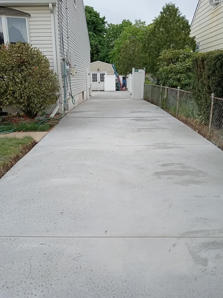 Concrete driveway between houses, bordered by shrubs and chain-link fence, leading to a garage.