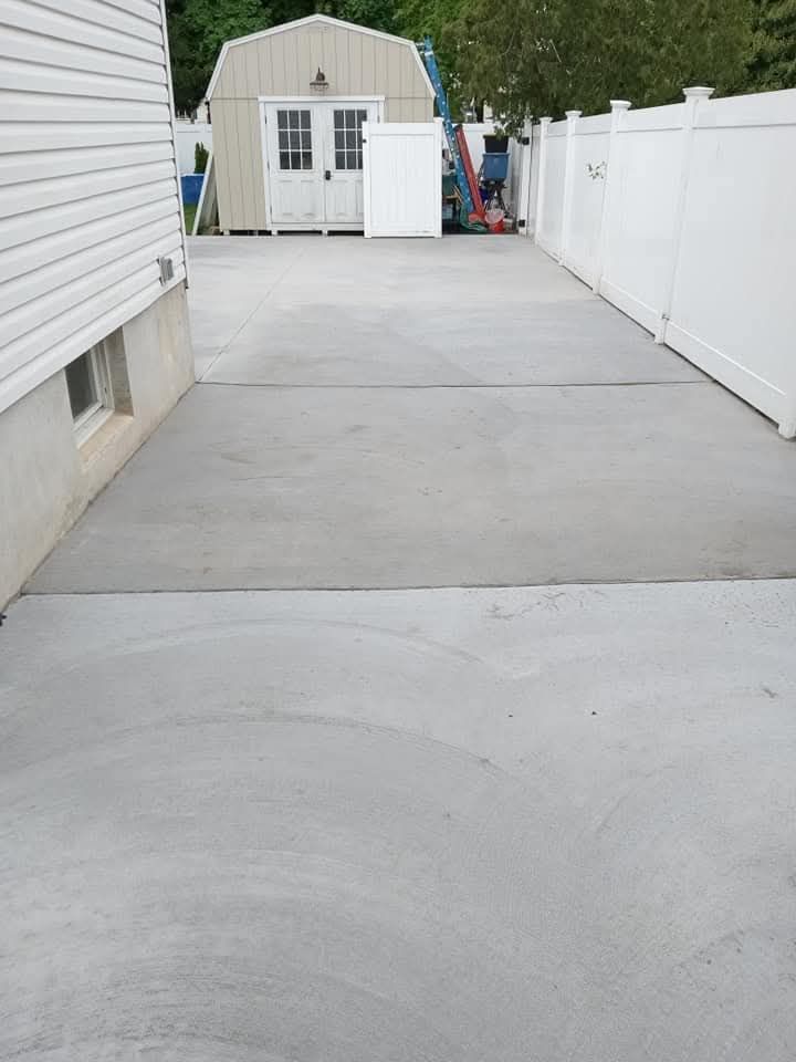 Concrete driveway between a house and white fence leading to a small shed at the end.