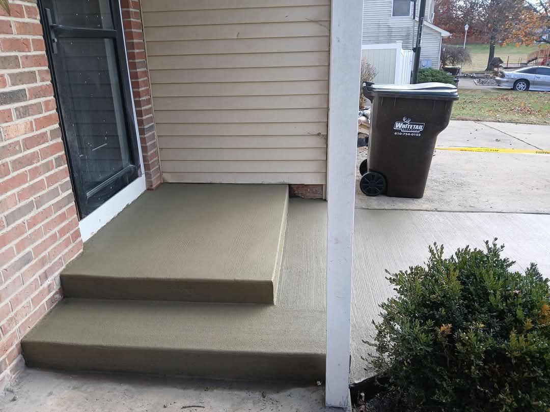Front porch with two concrete steps, a white post, brick wall, and trash bin by the driveway.