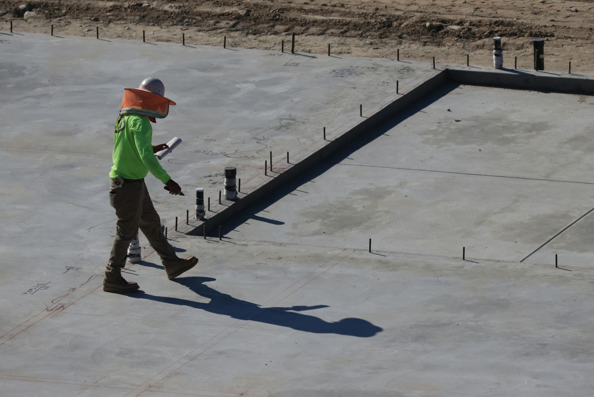 Construction worker in neon green jacket and orange hard hat walking on a concrete rooftop.