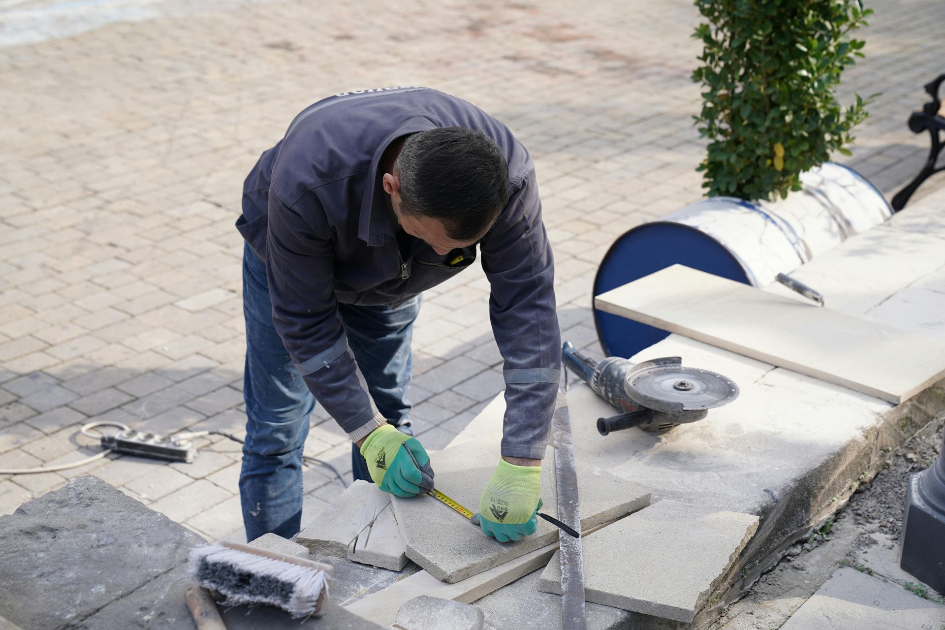 Worker in green gloves cutting concrete tiles with a saw on a paved sidewalk.