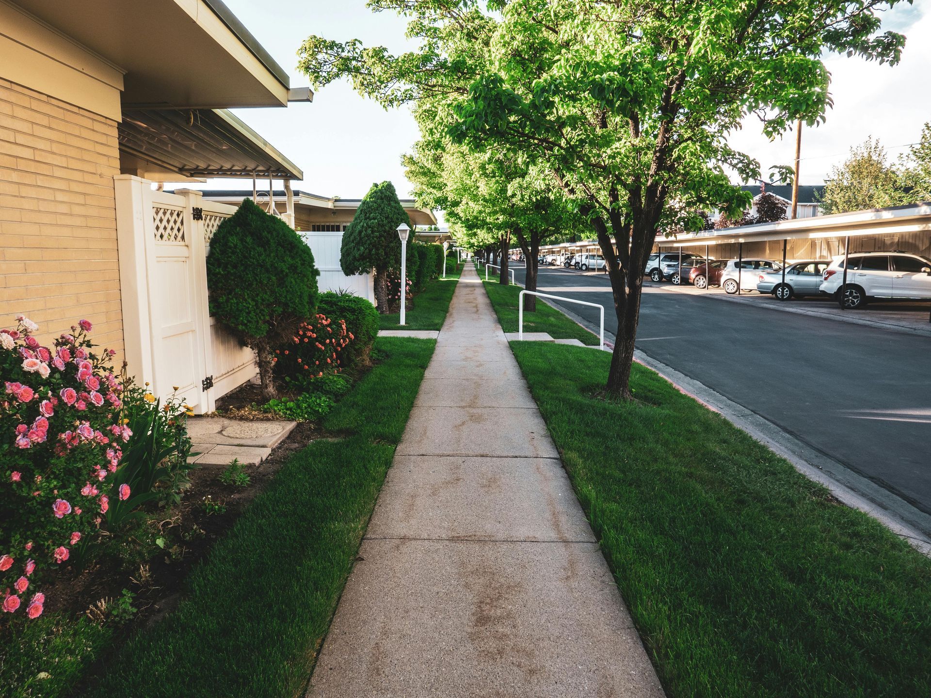Sidewalk beside a beige apartment building, with trees, flowers, and a parking lot on the right.