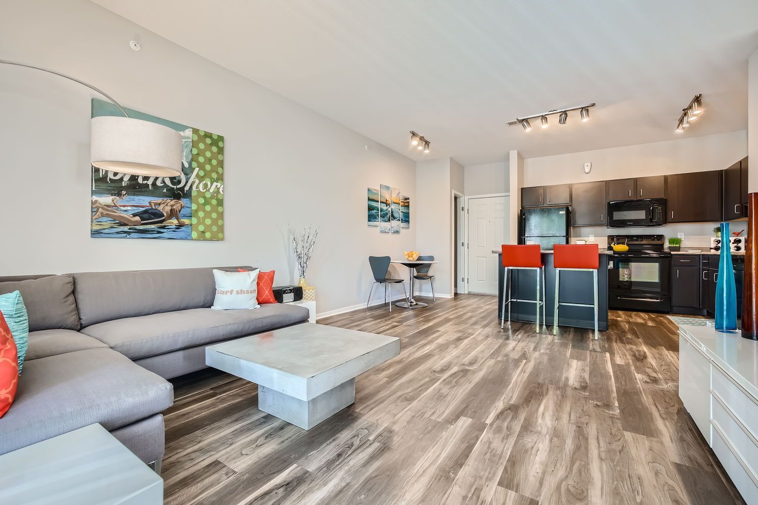 Open-concept living area with a gray sectional, wall art, and a dark kitchen with red bar stools.