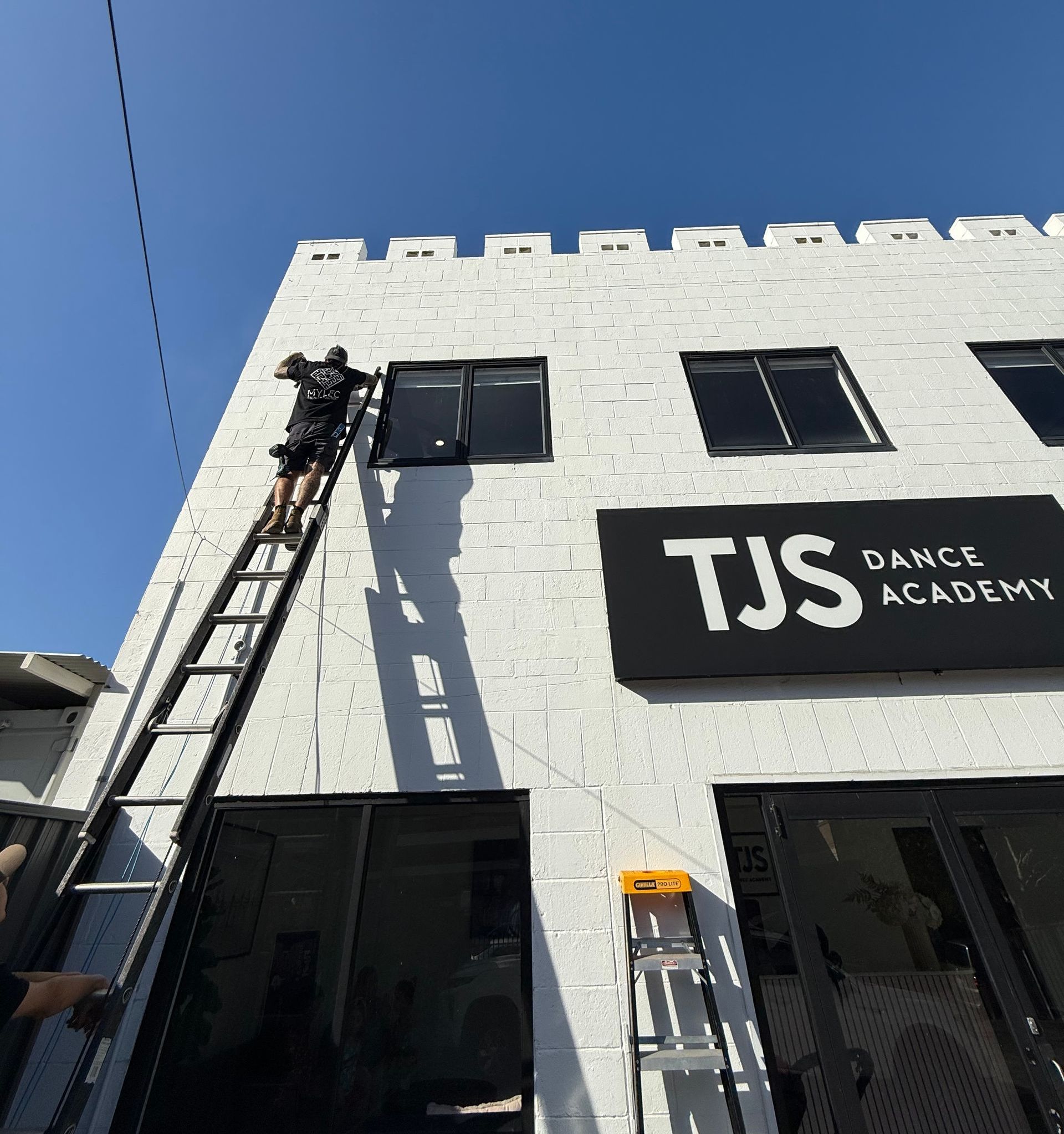 A man on a ladder in front of a building that says TJS Dance Academy — Mylec Electrical Solutions in Shell Cove, NSW