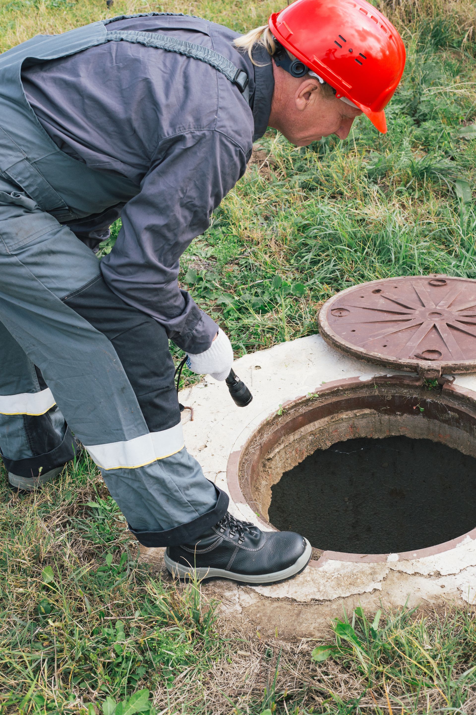 Worker in red hard hat inspecting an open manhole on grass