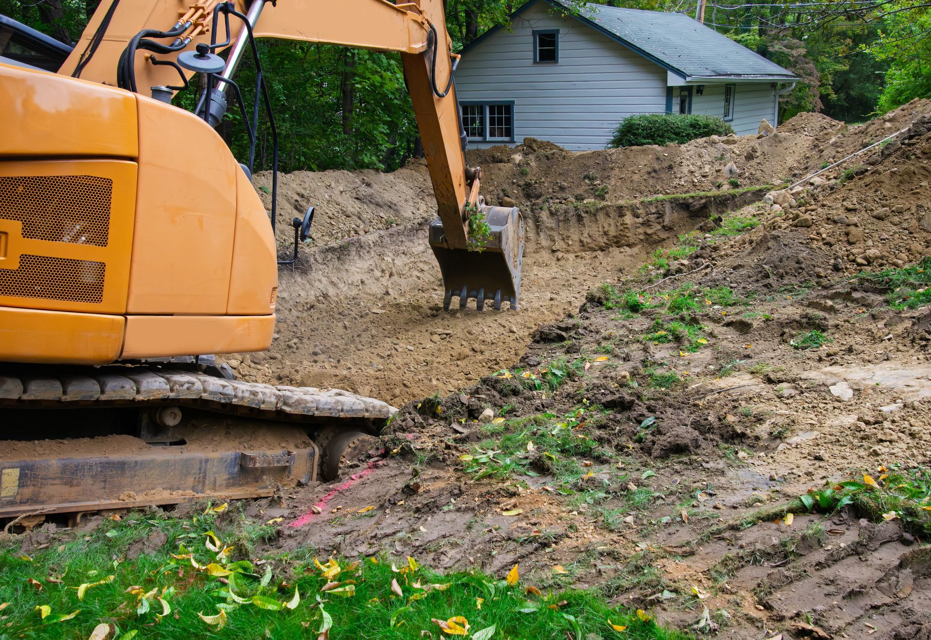 Yellow excavator digging dirt near a house on a muddy hillside construction site