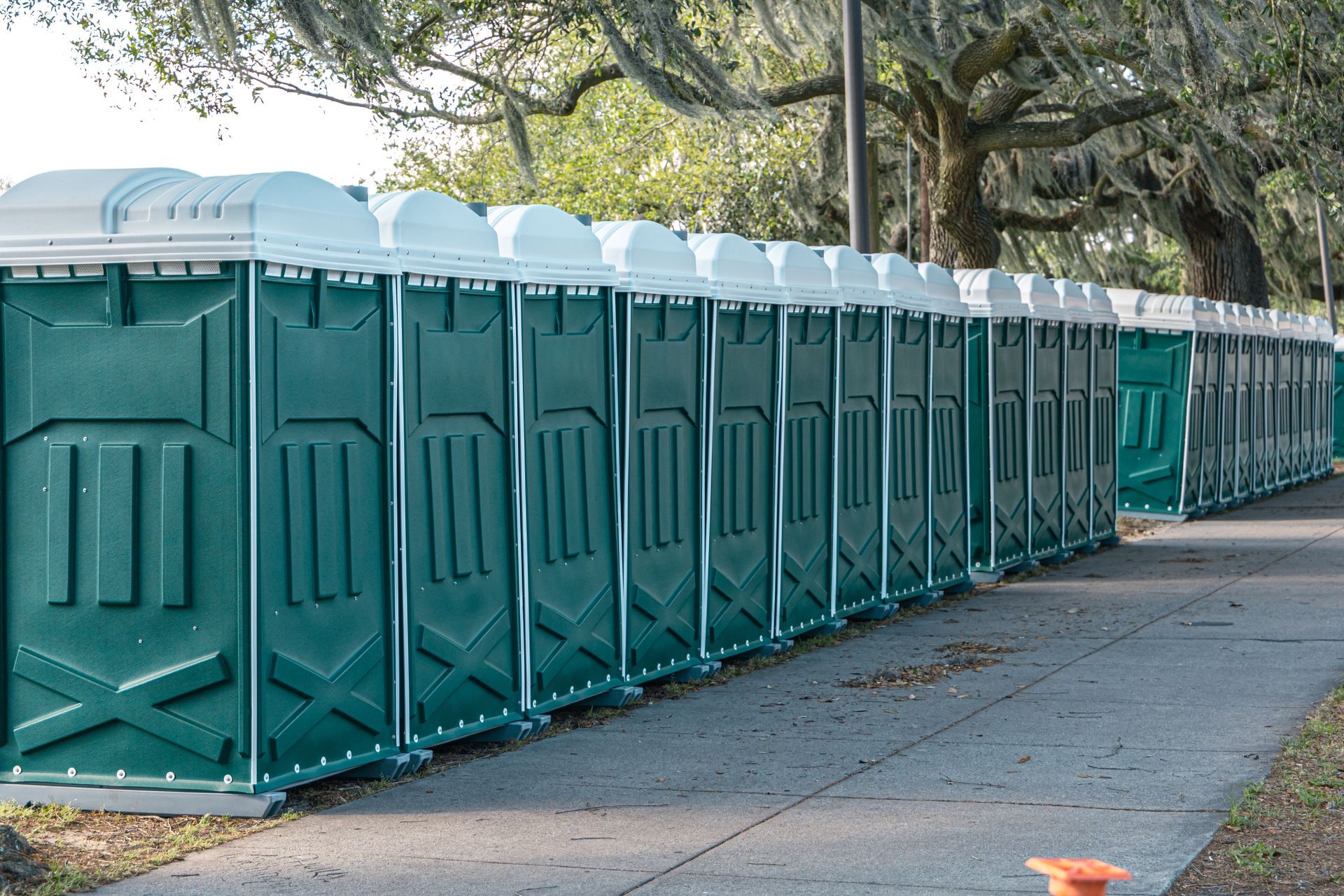 Row of green portable toilets along a tree-lined sidewalk in a park area Row of green portable toilets along a tree-lined sidewalk in a park area