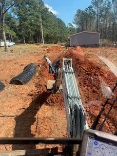 Trench with underground utility pipes and conduit at a rural construction site, with a shed and trees nearby