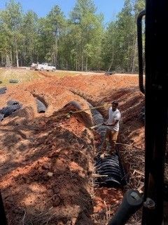 A man stands in a trench beside black pipes, with piles of orange dirt and trees in the background.