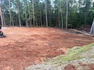 Cleared reddish dirt lot beside a forest, with a small tractor at left and grass in the foreground