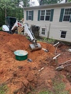 Backhoe excavating dirt beside a house with debris and a green container in the yard