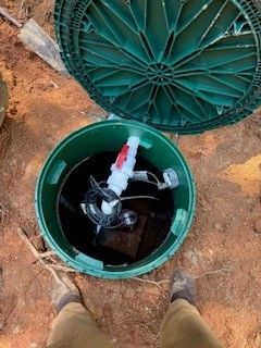 Open green utility access box in dirt with exposed pipes and valve inside, viewed from above