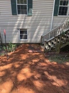 Backyard with red dirt ground, house siding, small basement window, and wooden stairs on the right