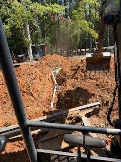 Excavator digging a large trench with exposed pipes in a wooded outdoor area