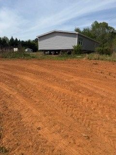 A white mobile home on blocks beside a reddish dirt lot under a clear sky.