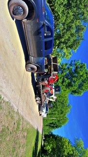 Blue pickup trucks parked on a dirt road with a red tractor, trees, and bright blue sky