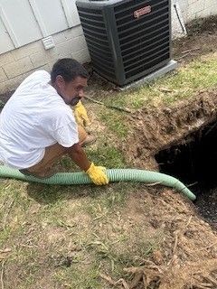 Worker in yellow gloves inserting a green hose into a large hole beside an HVAC unit.