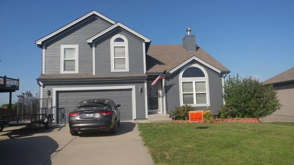 Suburban two-story gray house with a parked car in the driveway and an orange sign on the lawn.