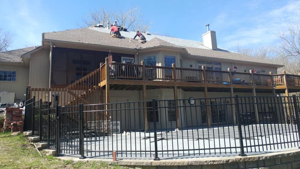 Large house with a wooden deck, black metal fence, and a person on the roof under a blue sky