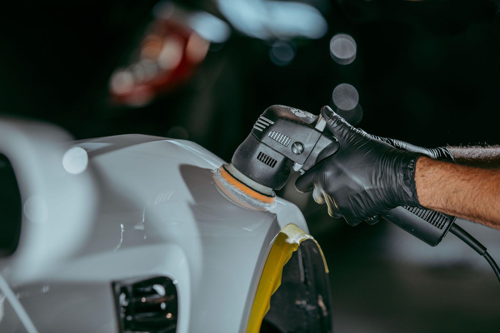 Gloved hand polishing a white car with a buffer in a dark garage