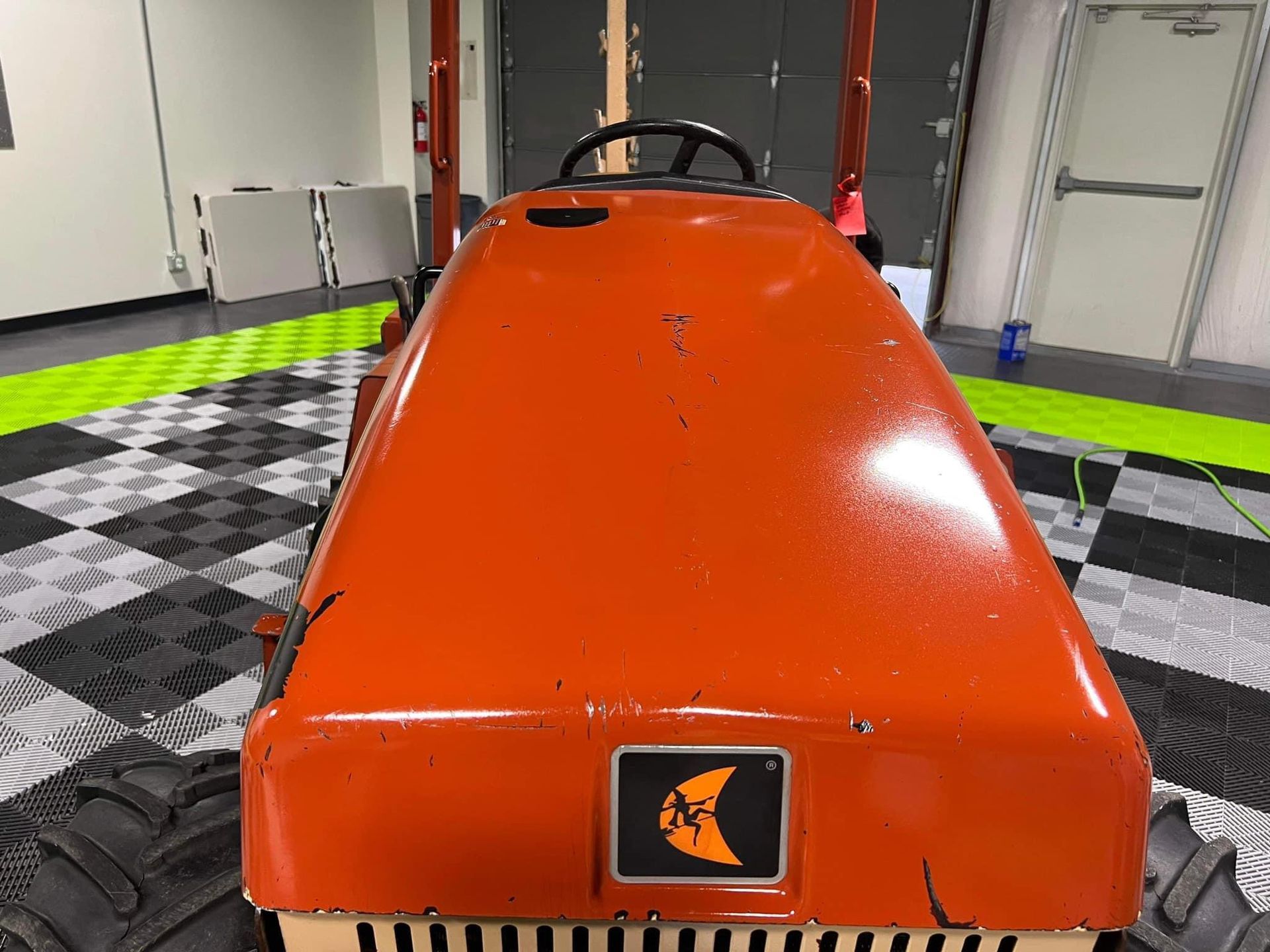Orange race car seen from above in a garage, with black tires and checkered floor.