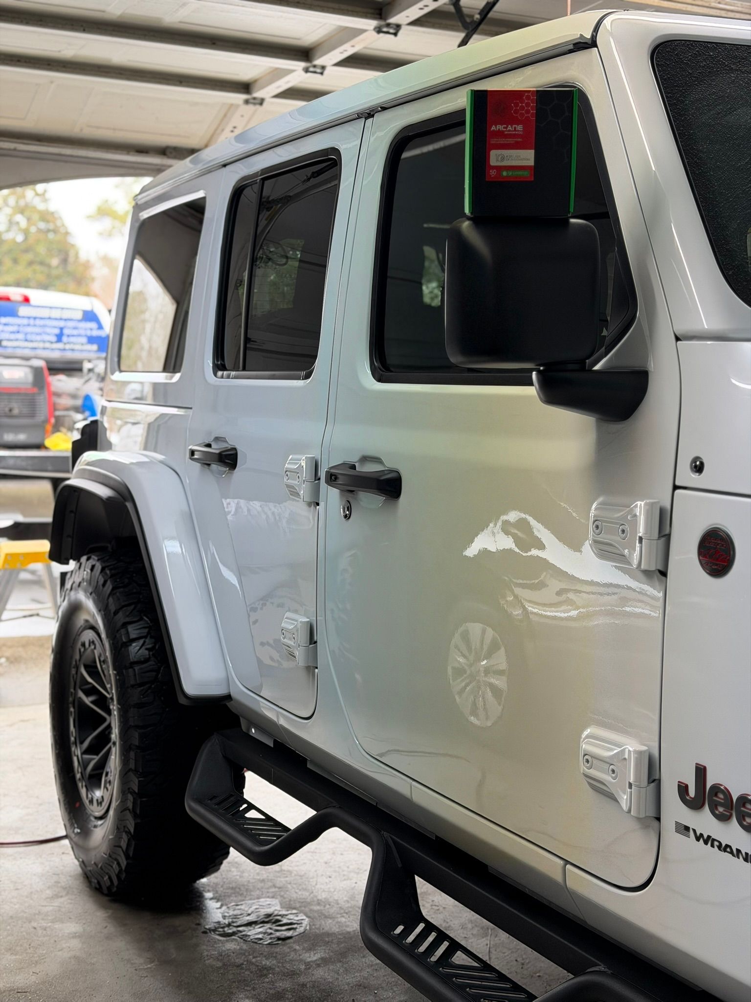 White Jeep Wrangler parked under a covered area, side view with black wheels and step rails.